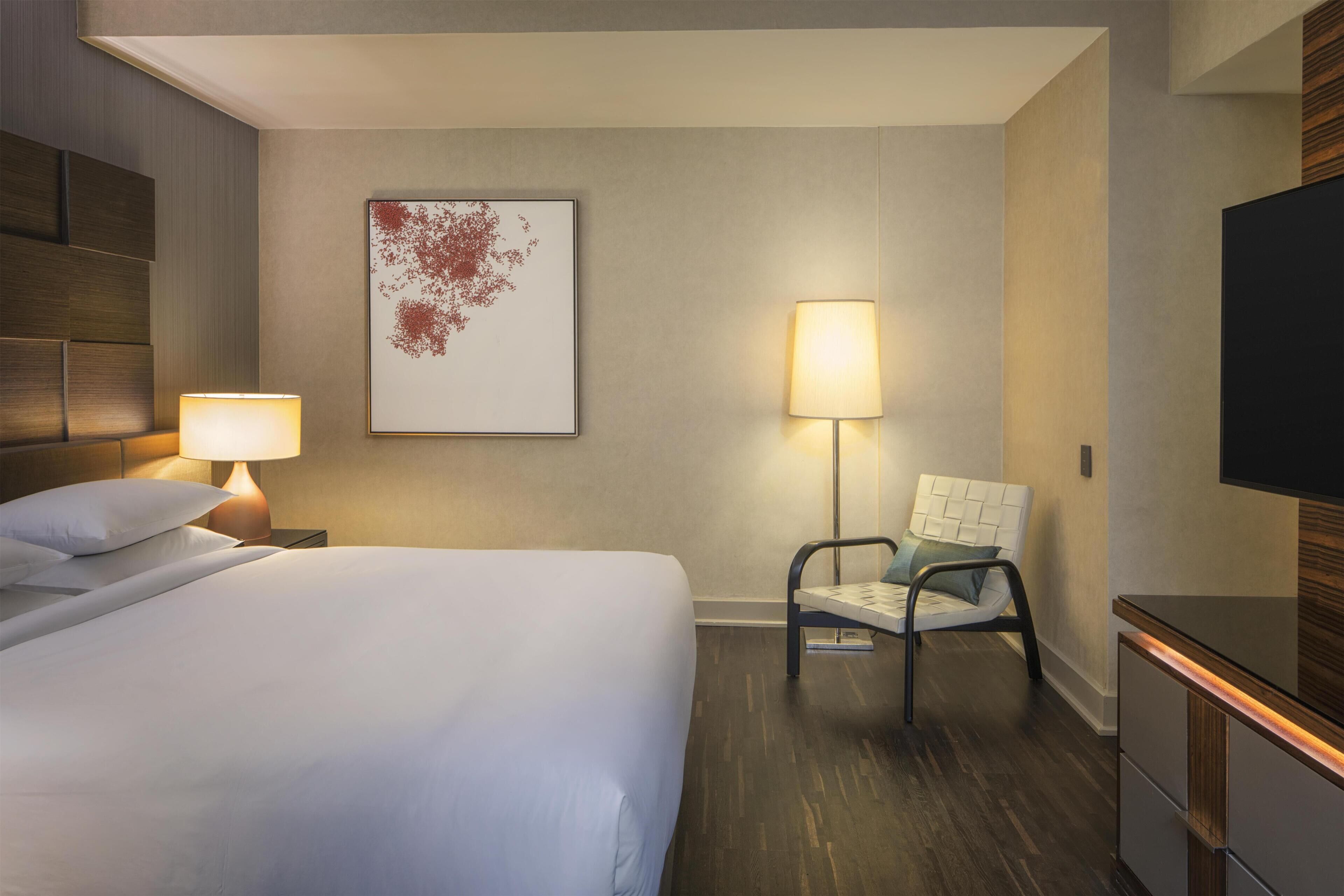 Habitación de hotel con cama, mesitas de noche, una silla, televisor y una obra de arte en la pared.