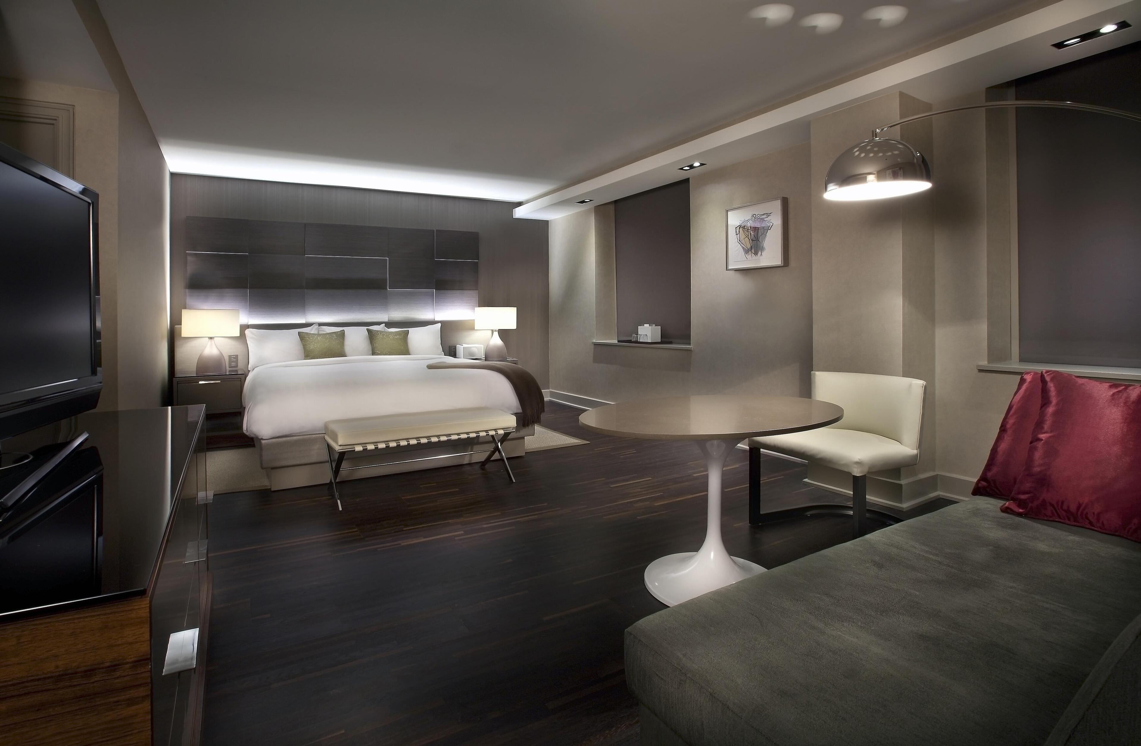 A modern hotel room with a bed, bedside tables, a TV, a round table, and a chair.