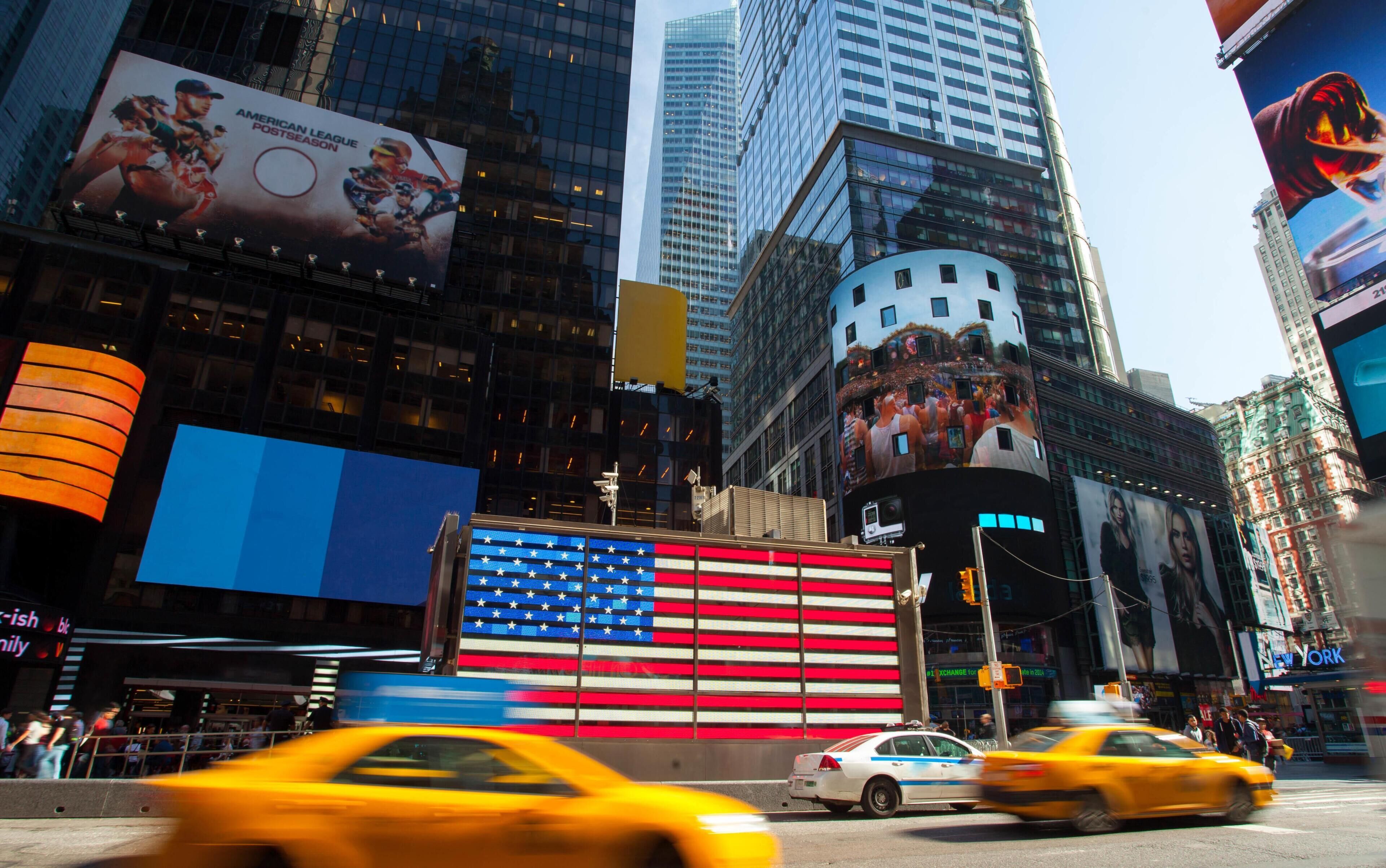 Uma rua urbana movimentada com edifícios altos, um mural grande com a bandeira americana e táxis amarelos.