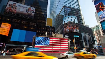 Uma rua urbana movimentada com edifícios altos, um mural grande com a bandeira americana e táxis amarelos.