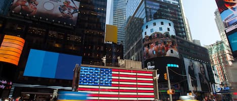 A busy urban street with tall buildings, a large American flag mural, and yellow taxis.