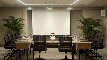 A conference room with a long table, black chairs, a projector screen, and two potted plants.