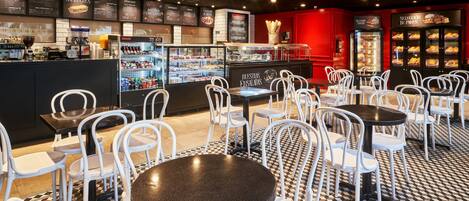 A café with white chairs and tables, a counter with a coffee machine, and a menu board displaying food items.