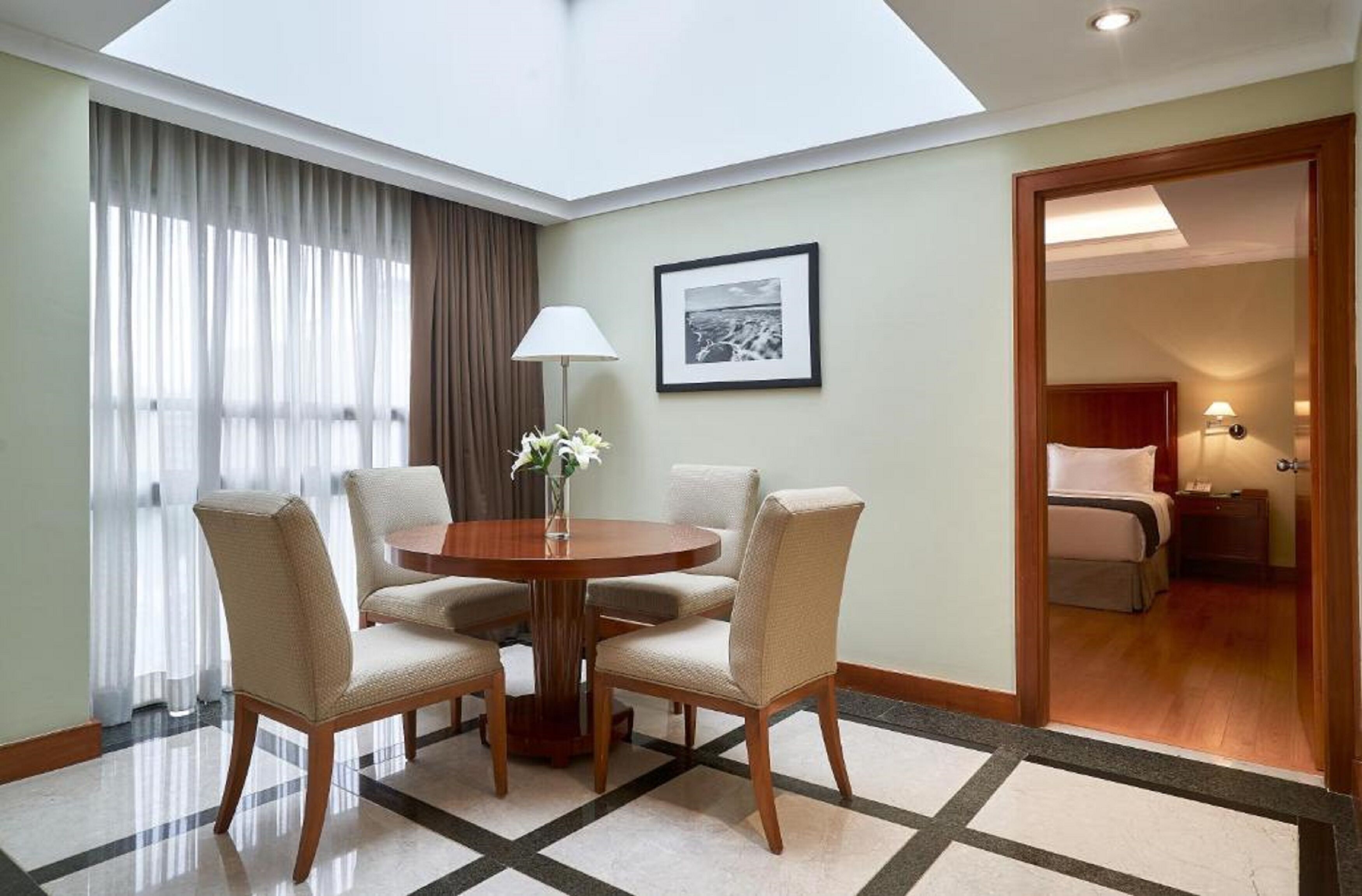 A hotel room with a round dining table, four chairs, a lamp, a framed picture on the wall, and a bed with a nightstand.
