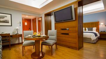 Habitación de hotel con piso de madera, una mesa redonda de comedor con dos sillas, un televisor montado en la pared, una cama con cabecera y una mesita de noche con lámpara.