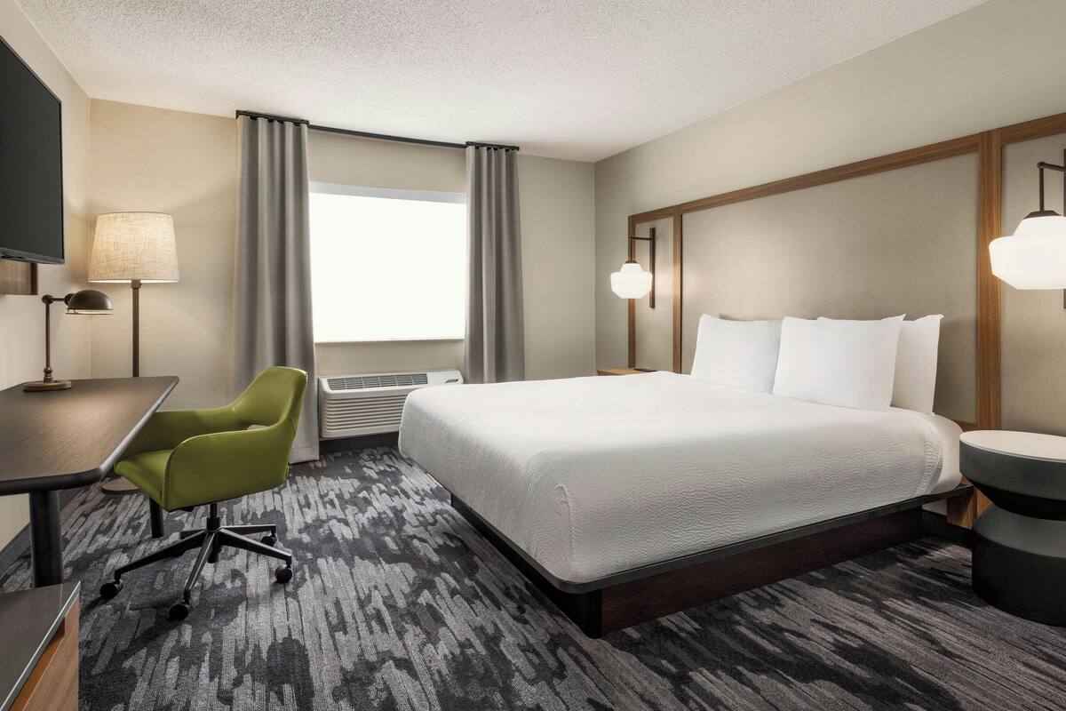 Une chambre d’hôtel avec un lit, un bureau, une chaise, une télévision et une fenêtre avec des rideaux.