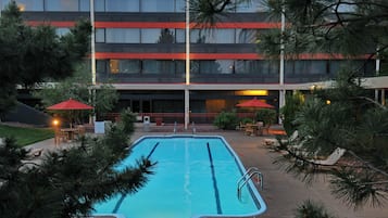 Una piscina con tabla de clavados, rodeada de árboles y un edificio de varios pisos y ventanas.