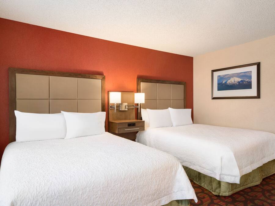Quarto de hotel com duas camas, uma mesa de cabeceira com abajur, um quadro na parede e um tapete vermelho.