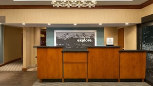 A reception desk with a wooden finish, a chandelier above, and a framed picture of mountains with the word "explore" on the wall. - Hampton Inn Denver West Federal Center (Lakewood)