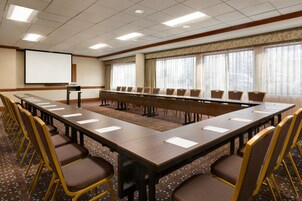 Un salón de conferencias con una larga mesa, sillas, una pantalla de proyector y ventanas grandes.