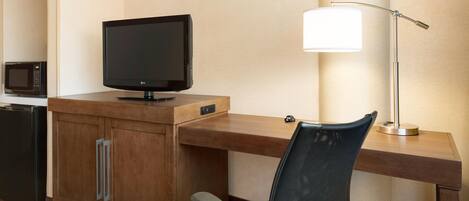 A hotel room with a wooden desk, a television, a lamp, a microwave, and a chair.