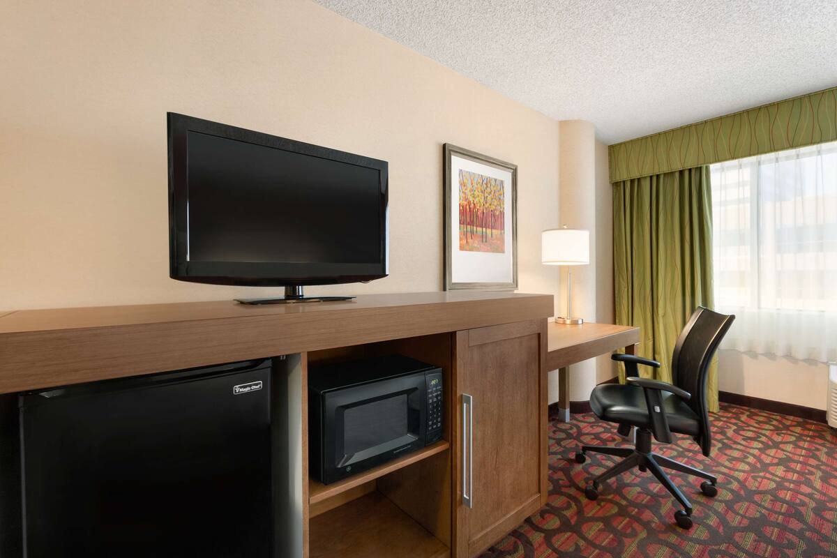 A hotel room with a flat-screen TV, a microwave, a refrigerator, a desk, a chair, a lamp, and a window with curtains.