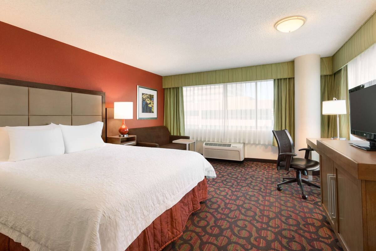 A hotel room with a large bed, a desk with a computer, a chair, a desk lamp, and a television.