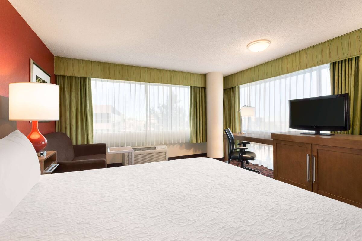 A hotel room with a large bed, a desk with a lamp, a television on a wooden cabinet, and a window with green curtains.