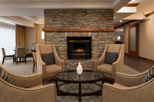 A hotel lobby with a stone fireplace, two armchairs, a round table, and a dining area in the background.