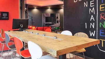 A modern meeting room with a long wooden table, colorful chairs, a blackboard with words, and a television.