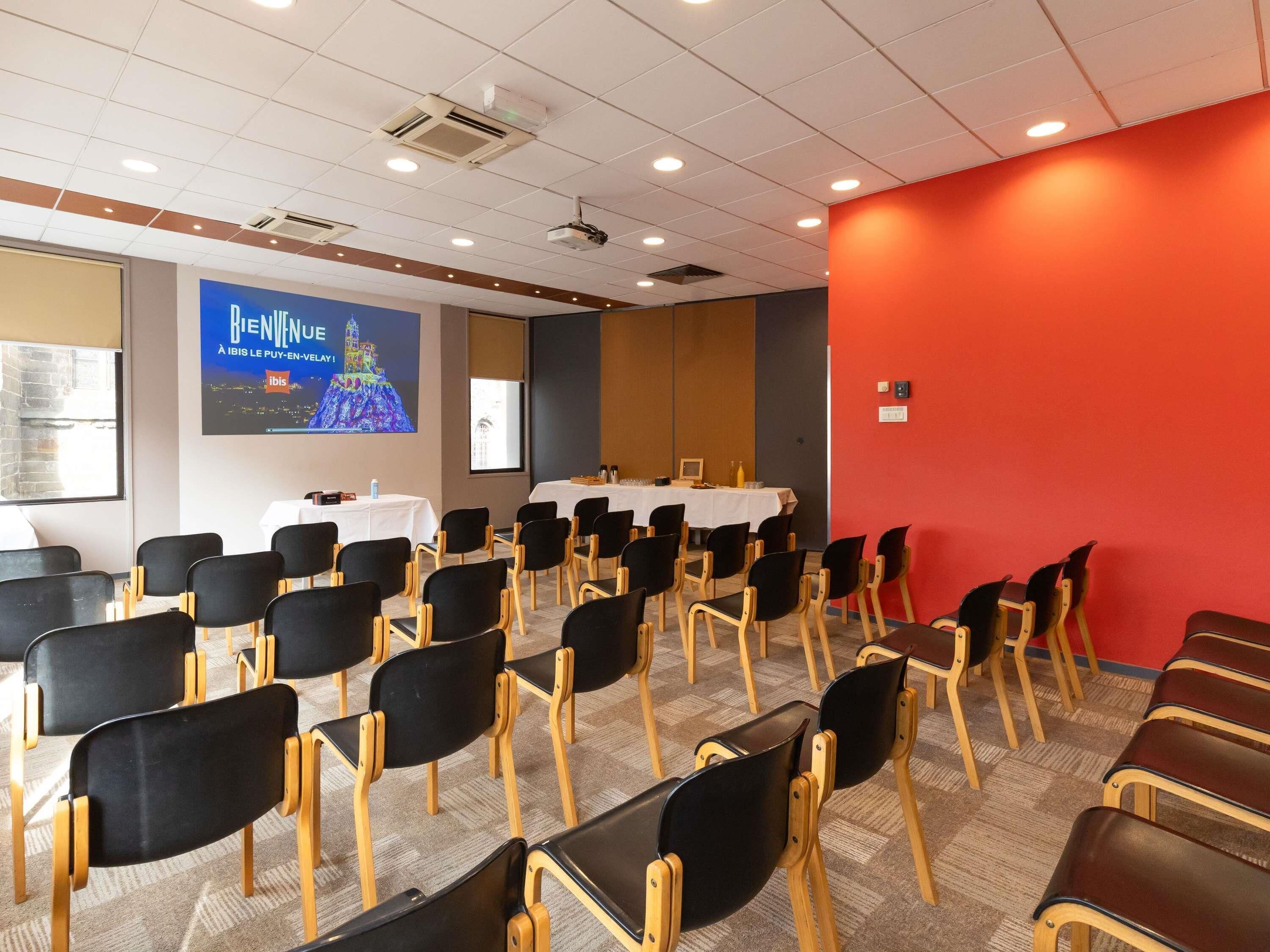 Un salón de conferencias con filas de sillas, una pantalla de proyector y una mesa cubierta con un mantel blanco.