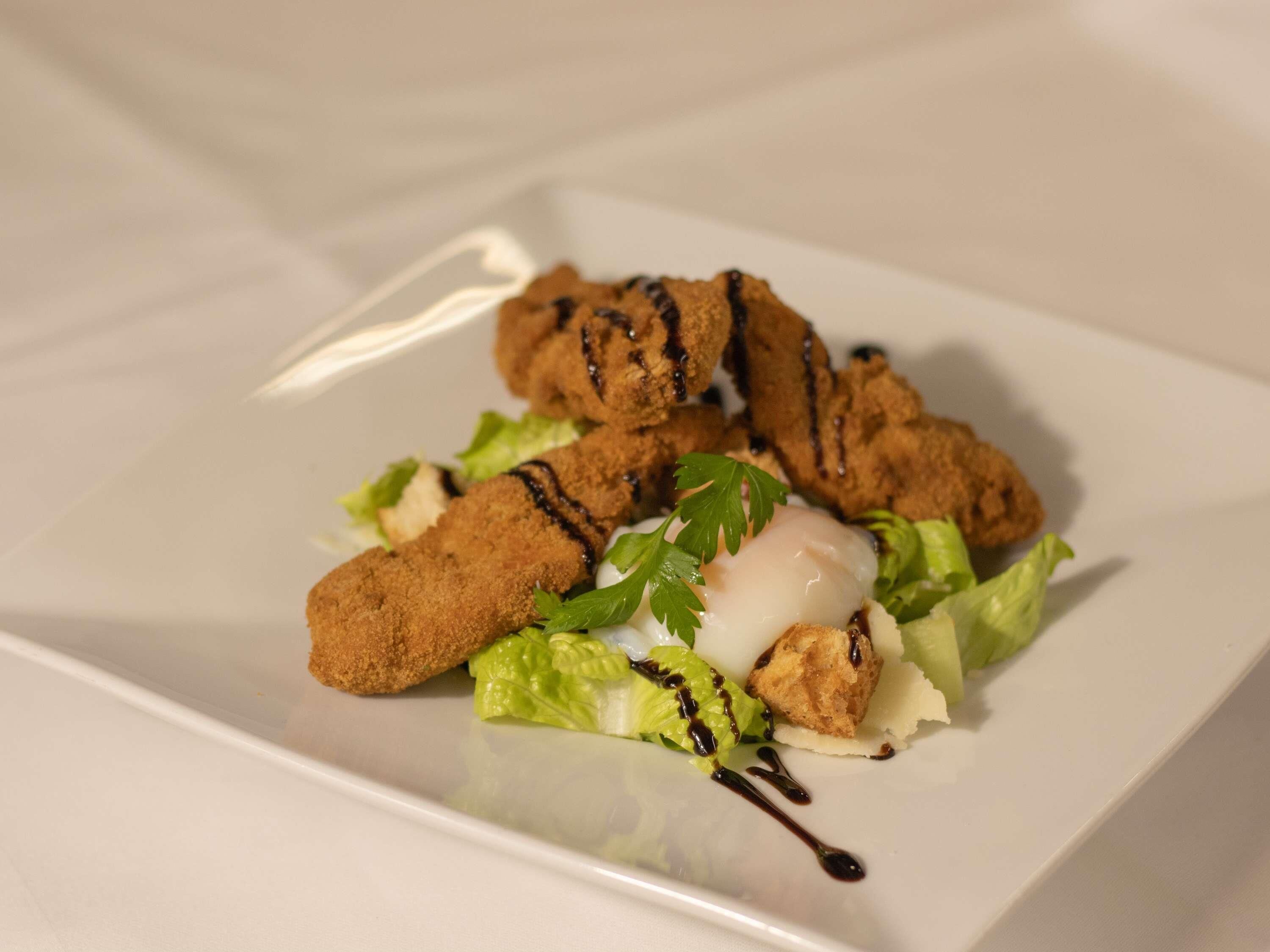 A square plate with a dish consisting of breaded items, possibly chicken, garnished with parsley and drizzled with a dark sauce.