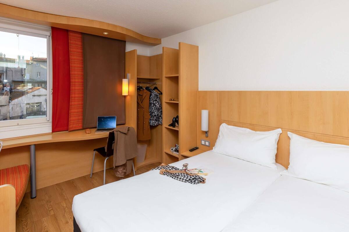 Camera d'albergo con un letto, una scrivania con un portatile, una sedia, un armadio e una finestra con tende.