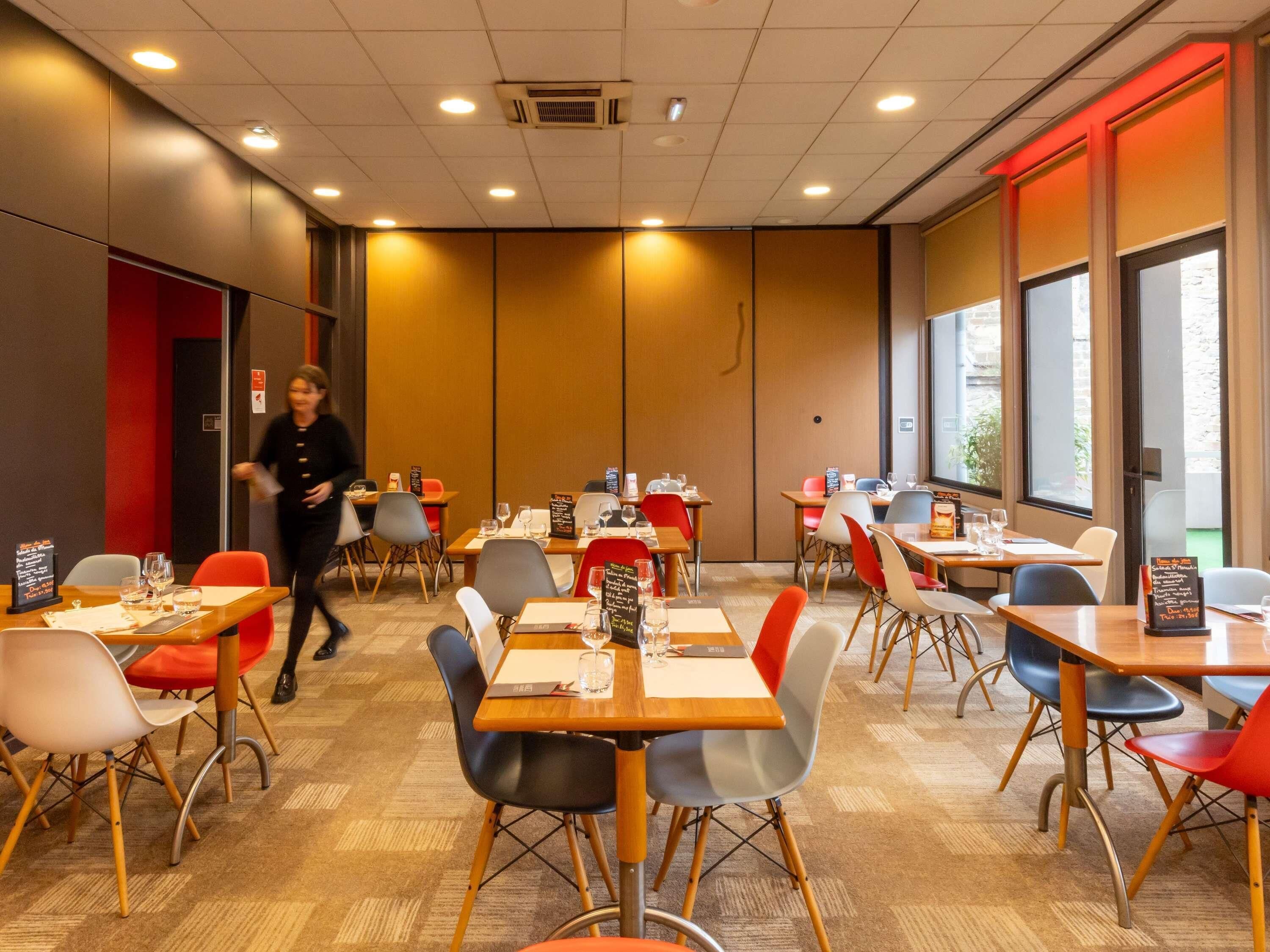 A modern dining area with multiple tables and chairs, a visible menu board, and a person walking in the background.