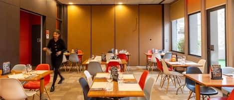 Uma área de refeições moderna com várias mesas e cadeiras, um quadro de menu visível e uma pessoa caminhando ao fundo.
