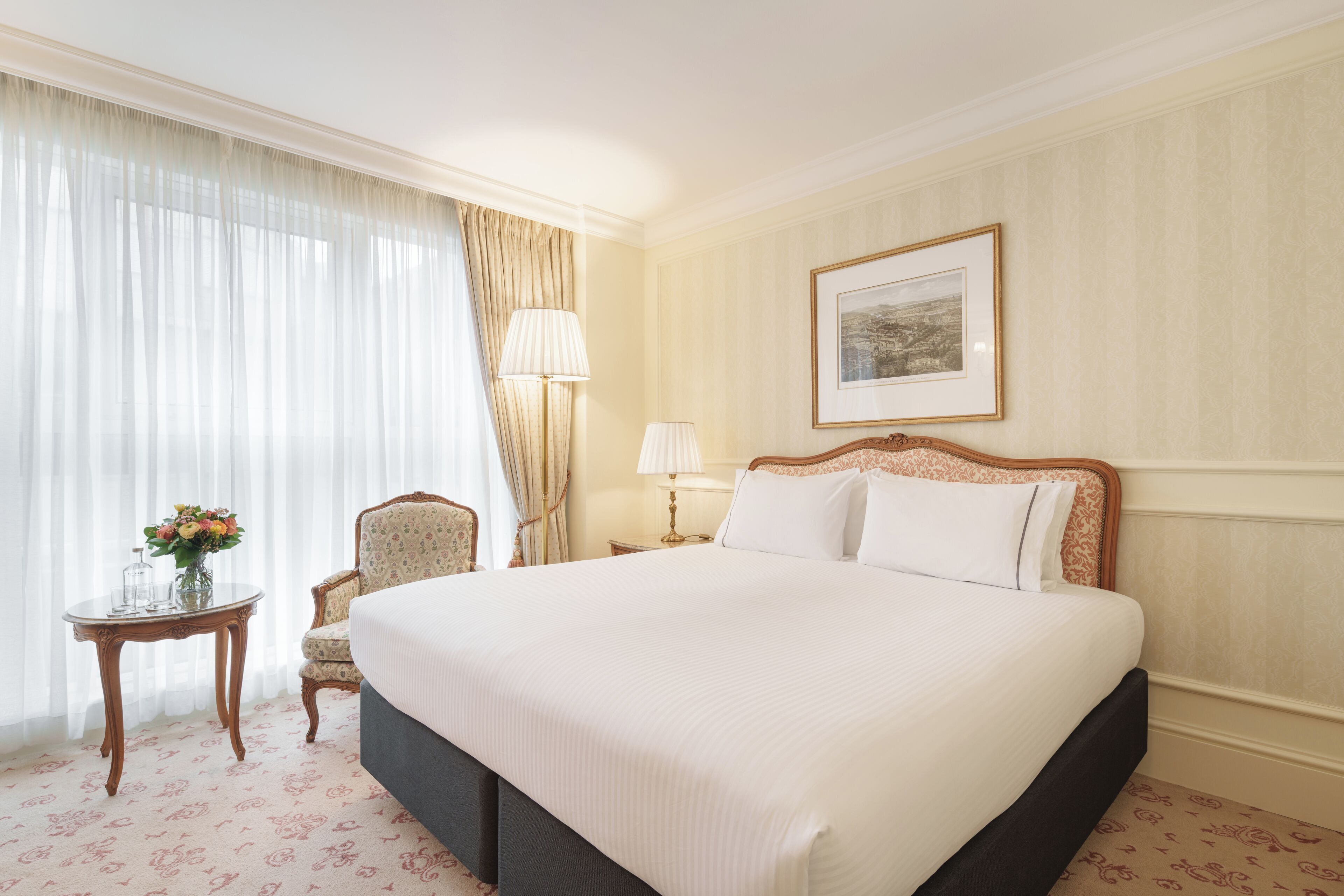 A hotel room with a large bed, a chair, a small table with a vase of flowers, and a framed picture on the wall.