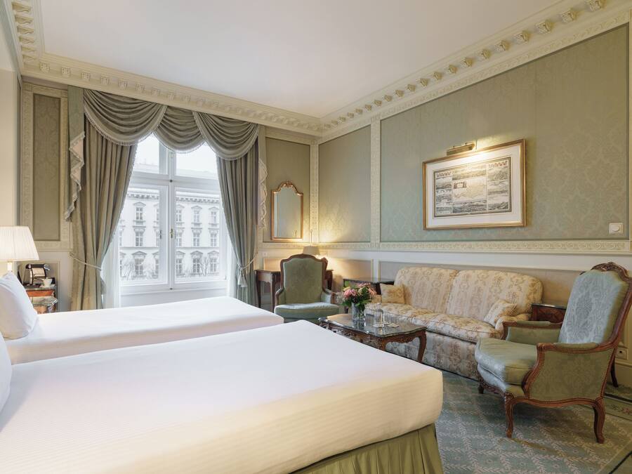 Habitación de hotel con dos camas, un sofá, una silla, una mesa con un florero y un cuadro en la pared.