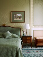 Quarto com cama, mesa de cabeceira com abajur, quadro pendurado na parede e pequena mesa com vaso e xícara.