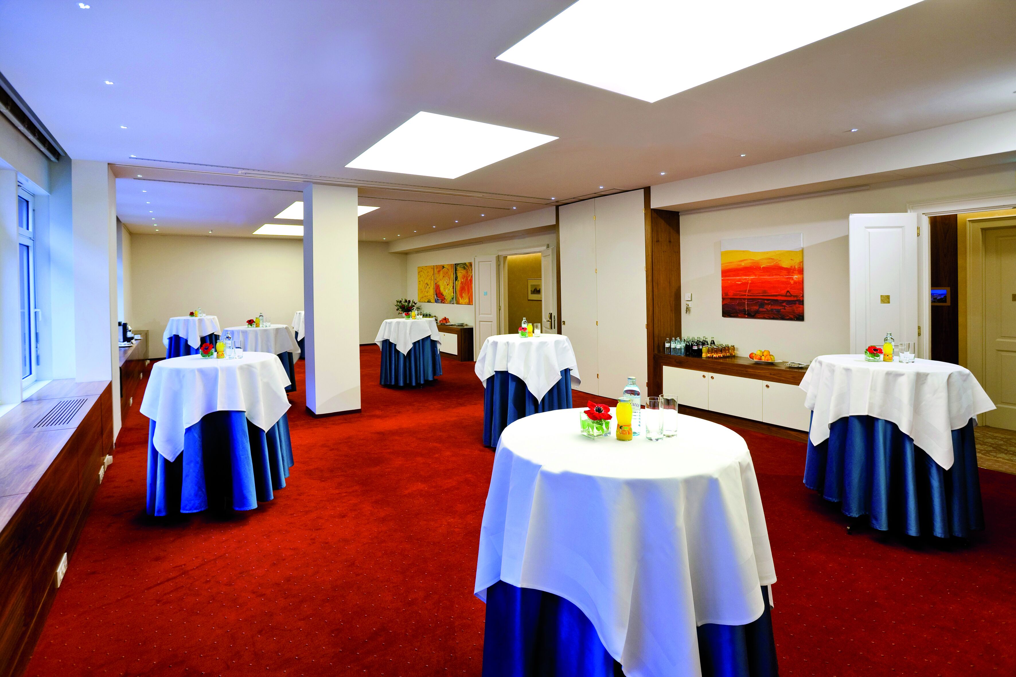 Habitación de hotel con alfombra roja, mesas blancas con manteles azules y un cuadro de atardecer en la pared.