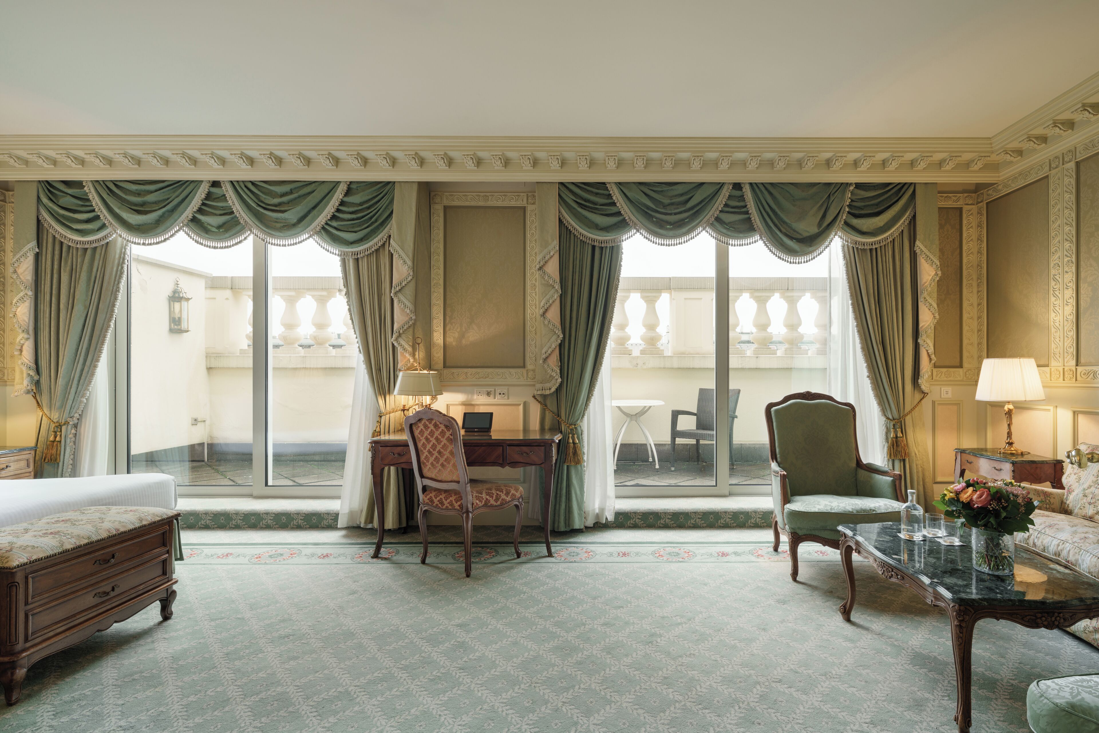A hotel room with a bed, a desk with a laptop, two chairs, a coffee table with a vase of flowers, and a balcony with a table and chairs.