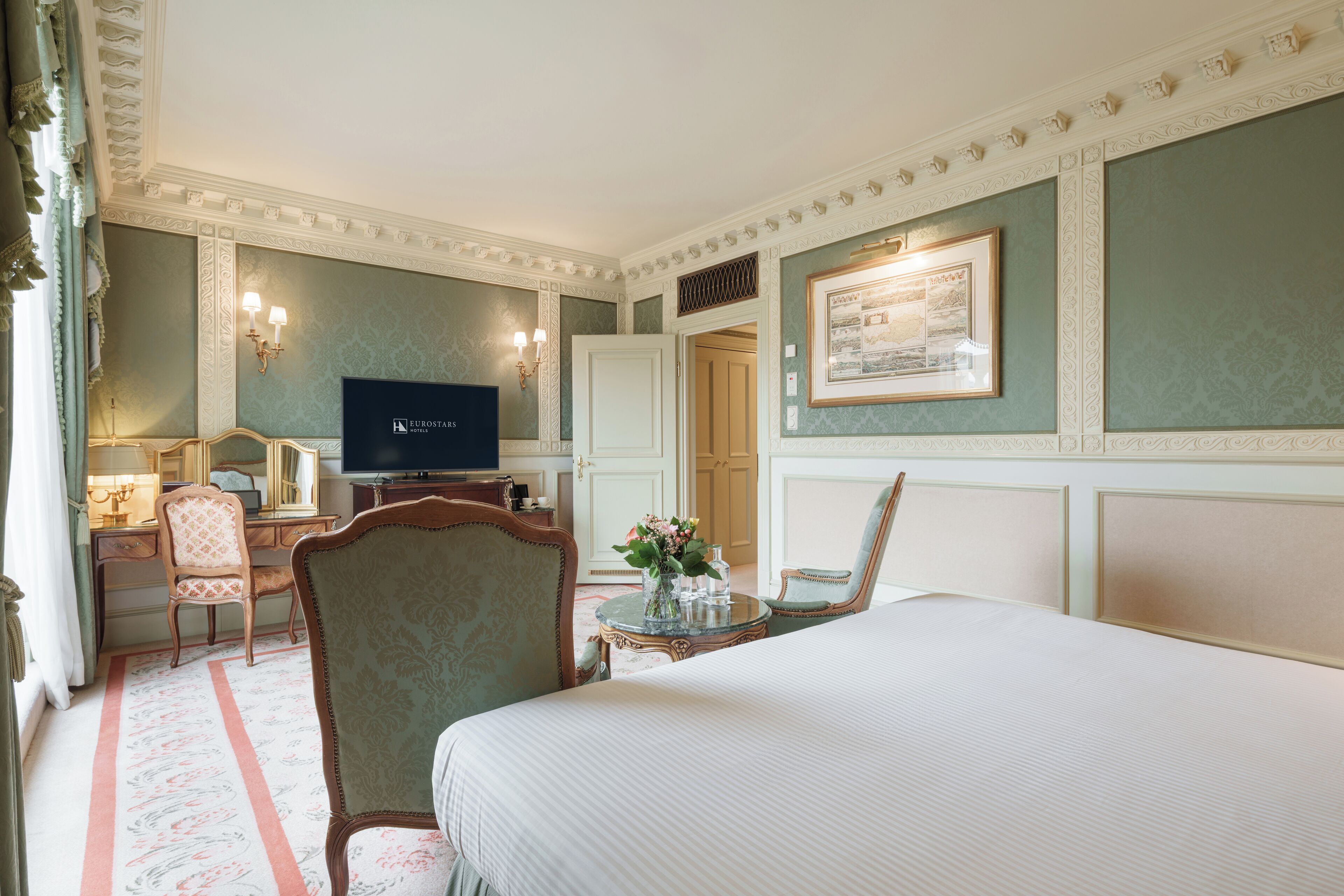 Een hotelkamer met een bed, een bureau met een televisie, een stoel en een klein tafeltje met een vaas.