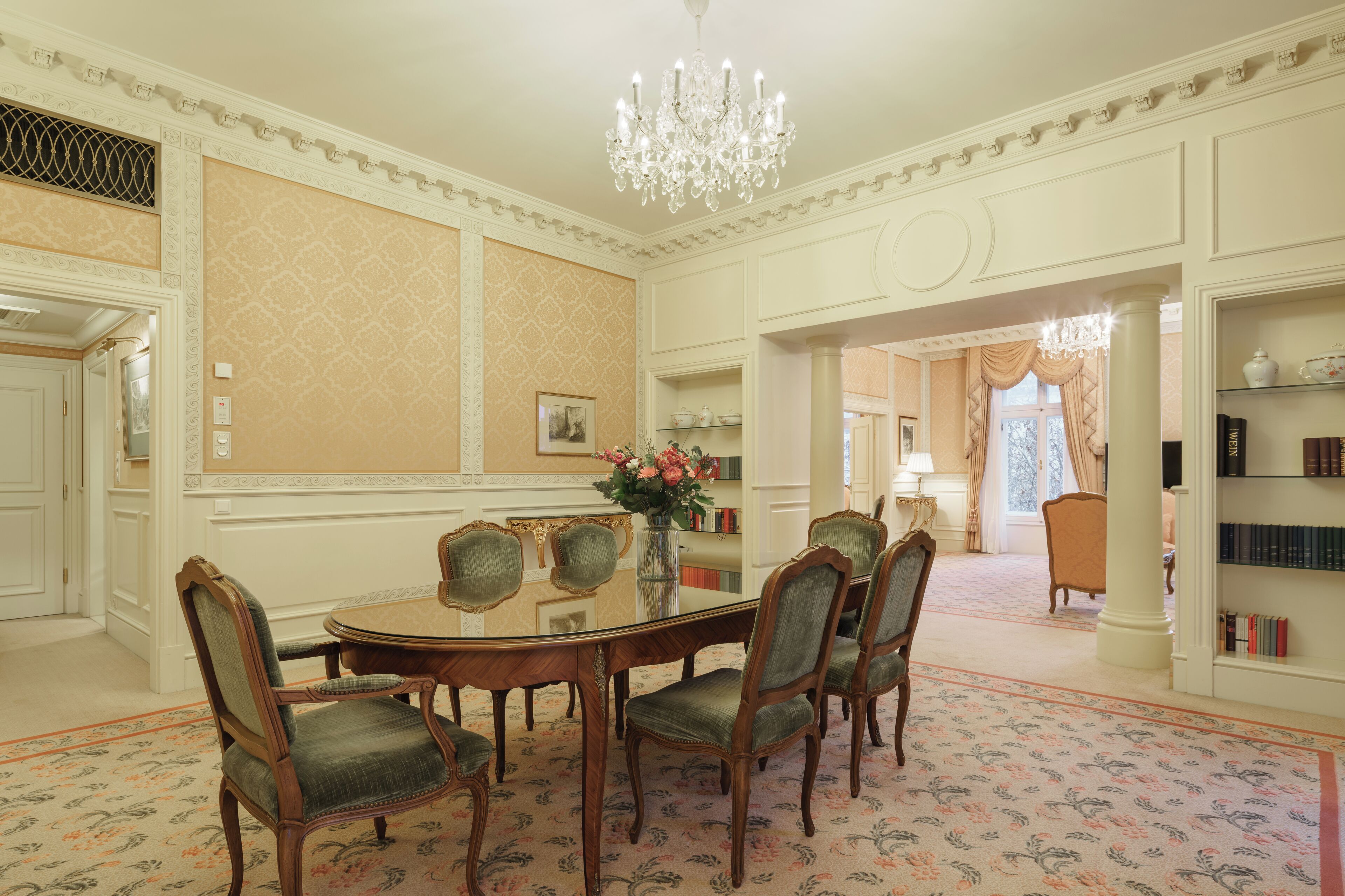 A formal dining room with a glass table surrounded by upholstered chairs, a chandelier, and a patterned carpet.