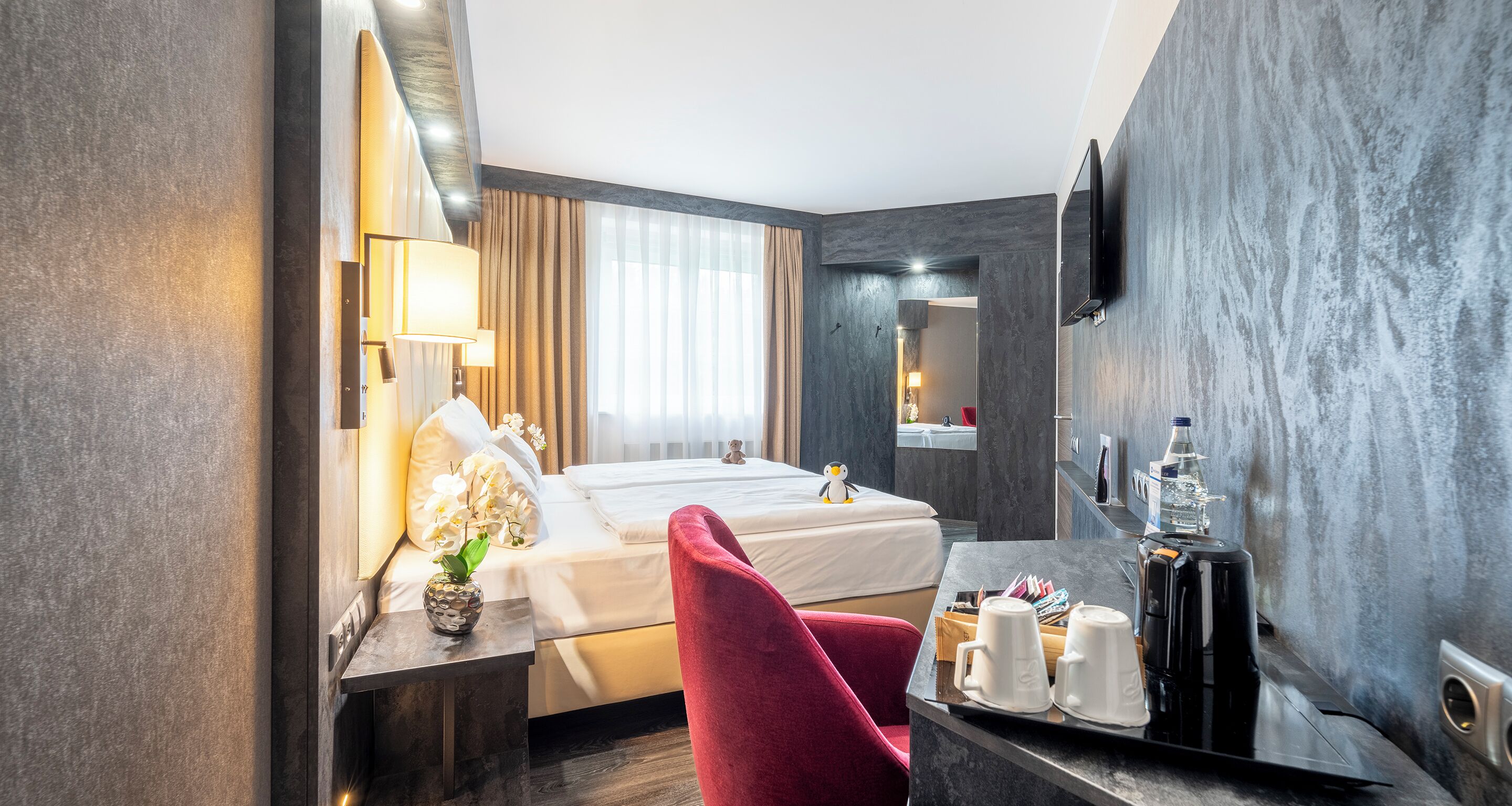 A hotel room with a bed, a desk with a lamp, a chair, a coffee maker, and a TV mounted on the wall.