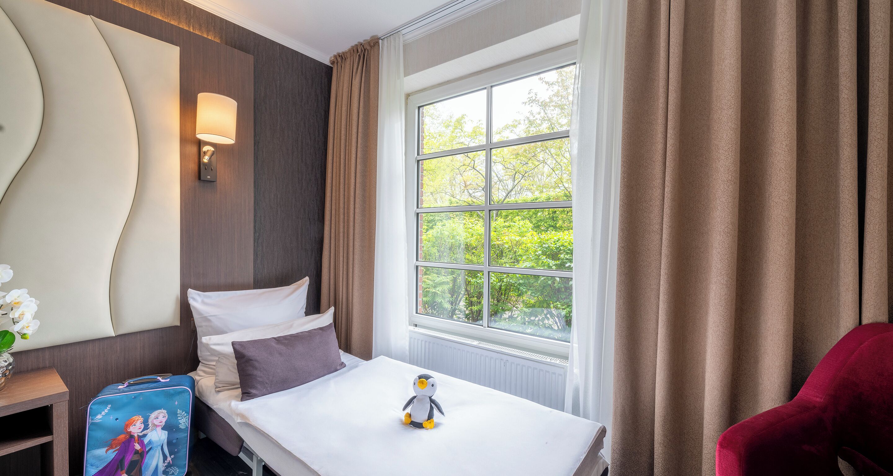 Ein Hotelzimmer mit einem Bett, einem Vorhangfenster, einer Wandlampe und einem Stofftier auf dem Bett.
