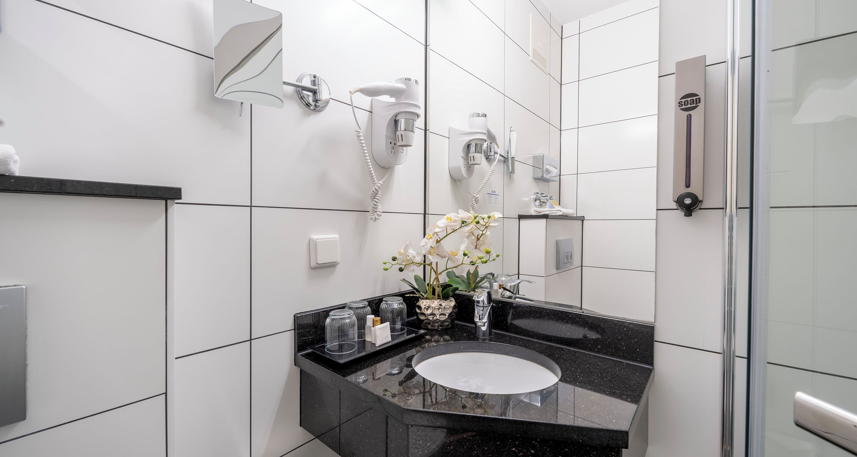 A modern bathroom with a black granite countertop, a white sink, and a mirror. There are two wall-mounted hair dryers and a towel rack.