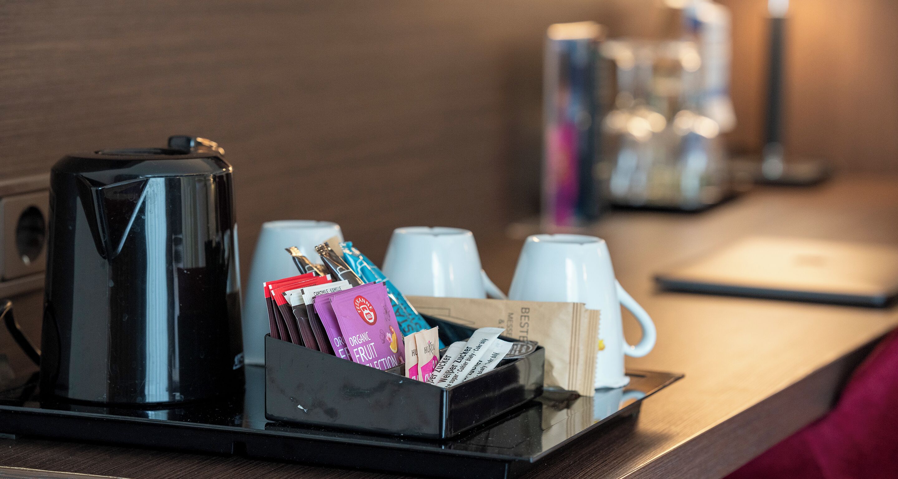 A coffee station with a coffee maker, cups, and a tray with coffee packets and a sugar packet.