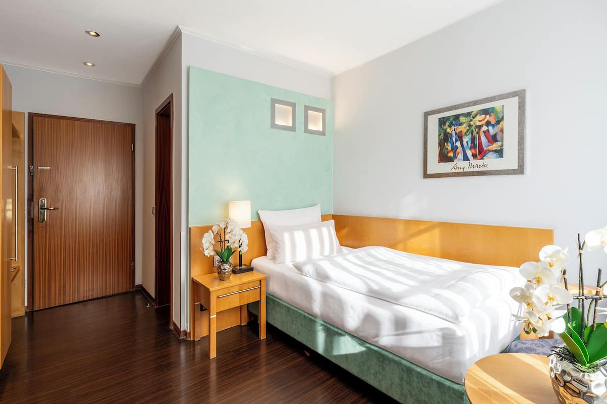 Quarto de hotel com uma cama, mesa de cabeceira com abajur, um vaso de flores e um quadro na parede.