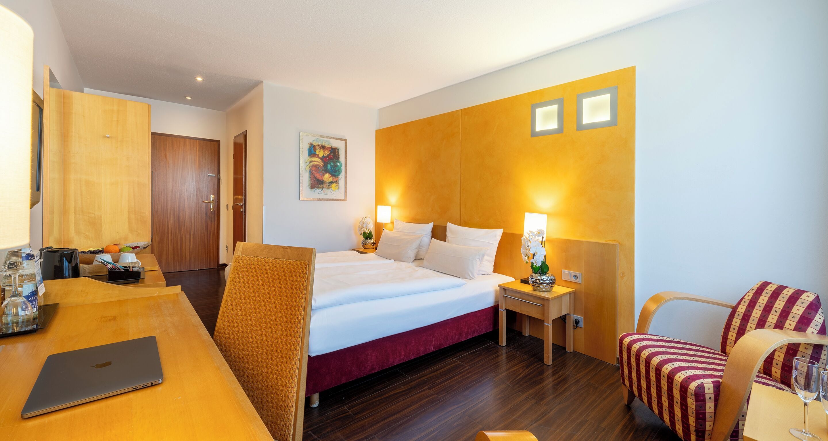 Ein Hotelzimmer mit einem Bett, einem Schreibtisch mit Laptop, einem Stuhl und einem Nachttisch mit Lampe.