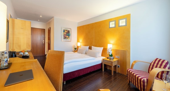 Camera d'albergo con un letto, una scrivania con un portatile, una sedia e un comodino con una lampada.