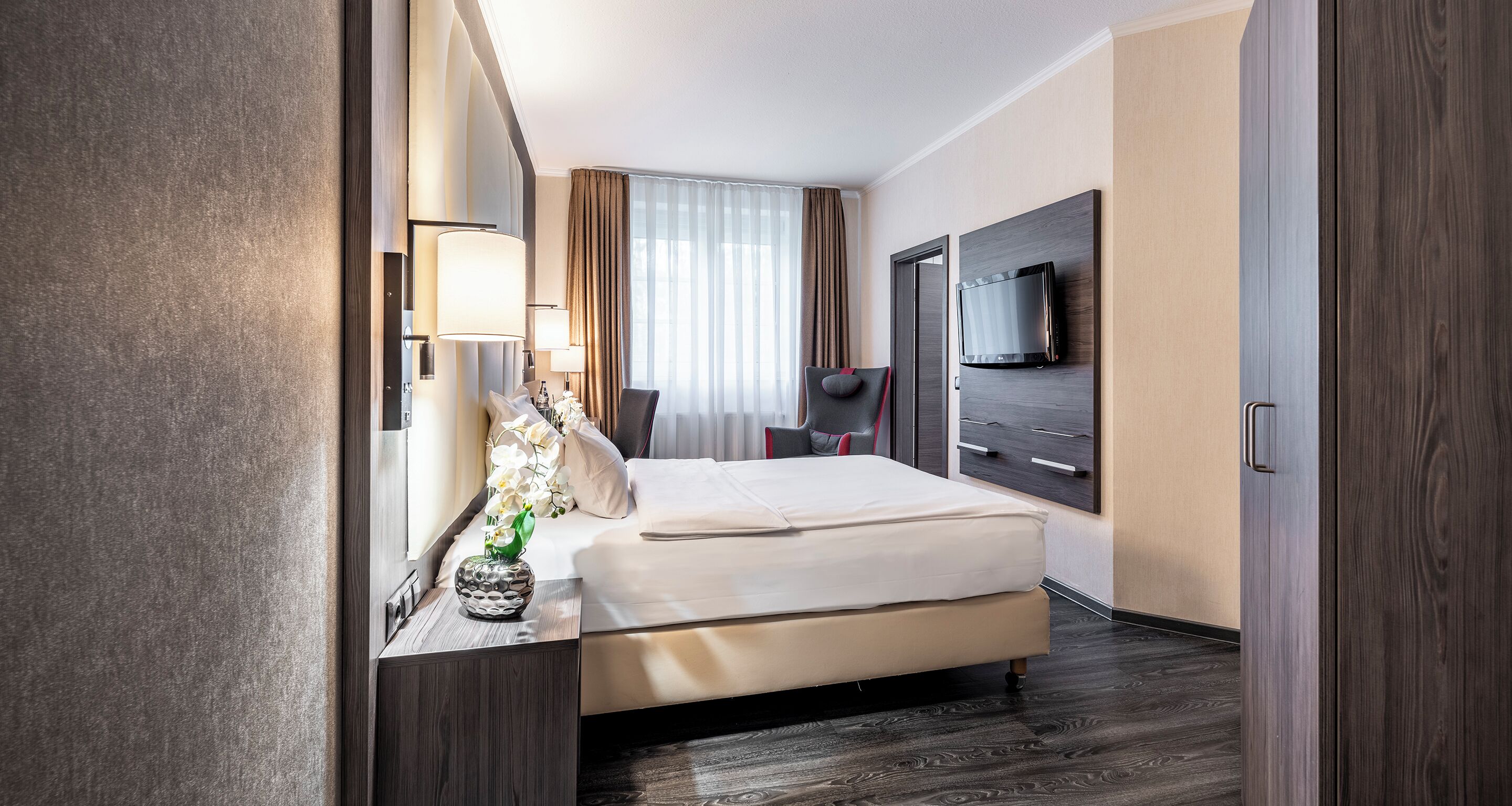 Ein Hotelzimmer mit einem Bett, Nachttischlampen, einem an der Wand befestigten Fernseher und einem Fenster mit Vorhängen.