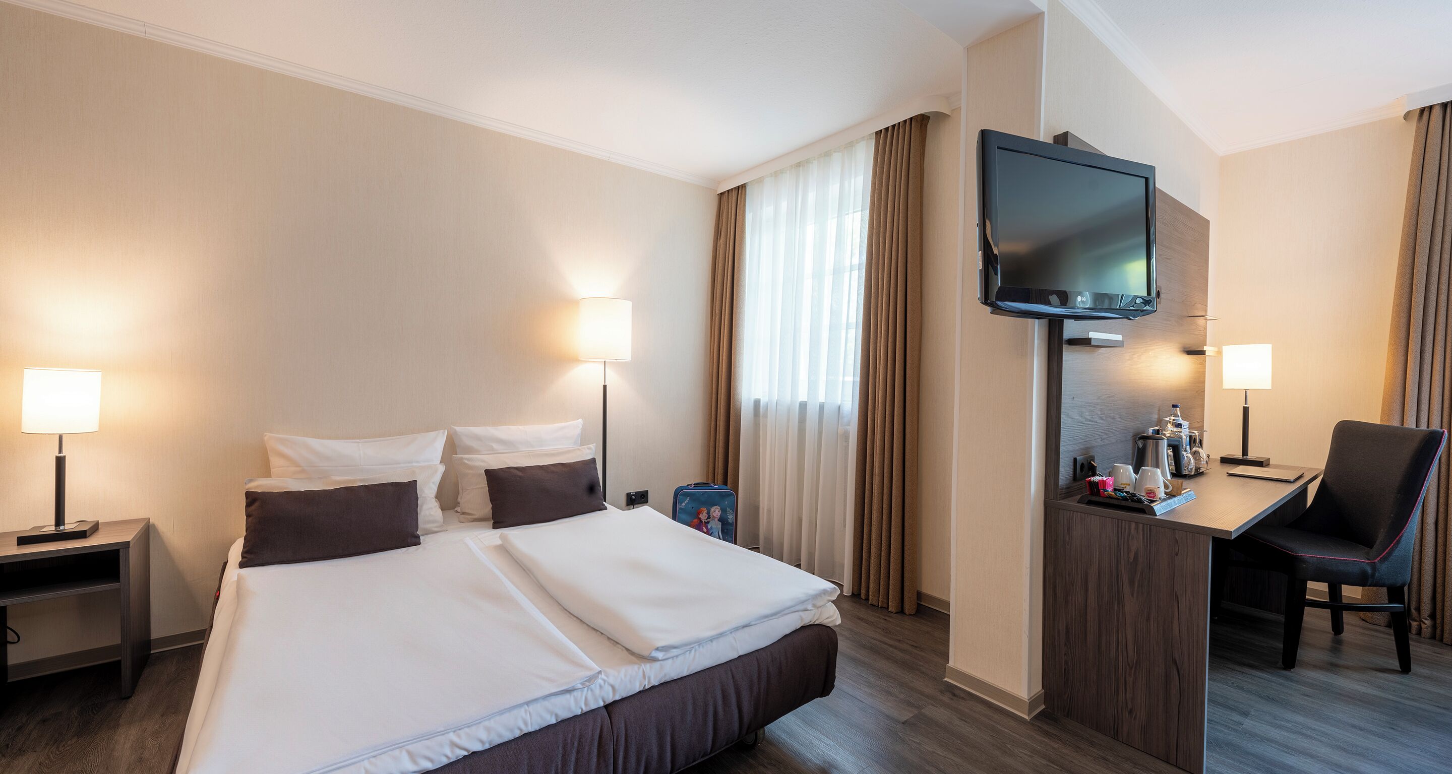 Ein Hotelzimmer mit einem großen Bett, einem Schreibtisch mit Stuhl, einem an der Wand montierten Fernseher und einem Fenster mit Vorhängen.