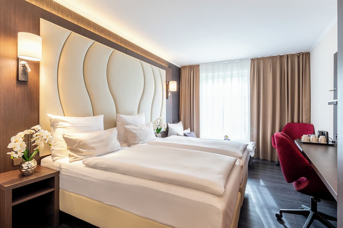 Quarto de hotel com duas camas, uma escrivaninha, uma cadeira e um vaso de flores.