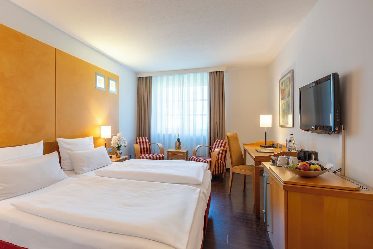 Quarto de hotel com duas camas, uma escrivaninha com cadeira, televisão e janela com cortinas.