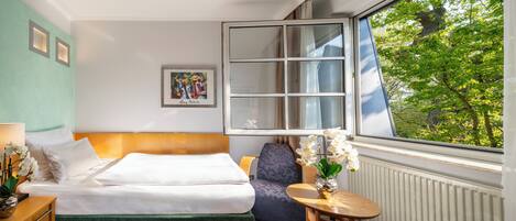Una habitación de hotel con una cama, una silla, una mesita con un florero y una ventana que da a árboles, además de un cuadro en la pared.