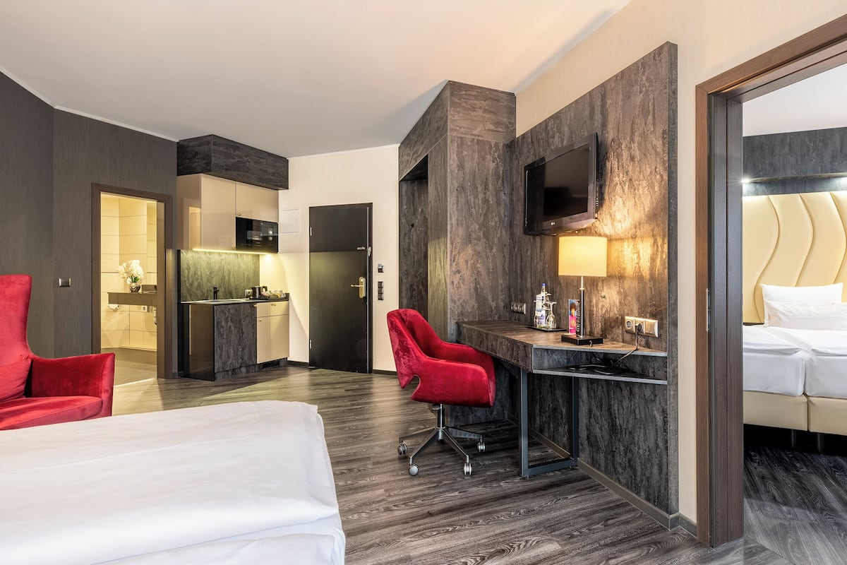 Quarto de hotel com cama, escrivaninha, uma cadeira vermelha, televisão e banheiro visível através de uma porta aberta.