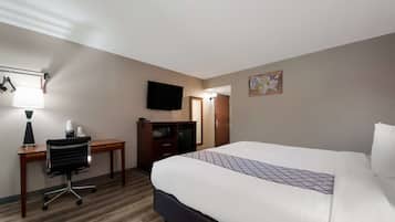A hotel room with a bed, desk, chair, TV, and a small table with a lamp.