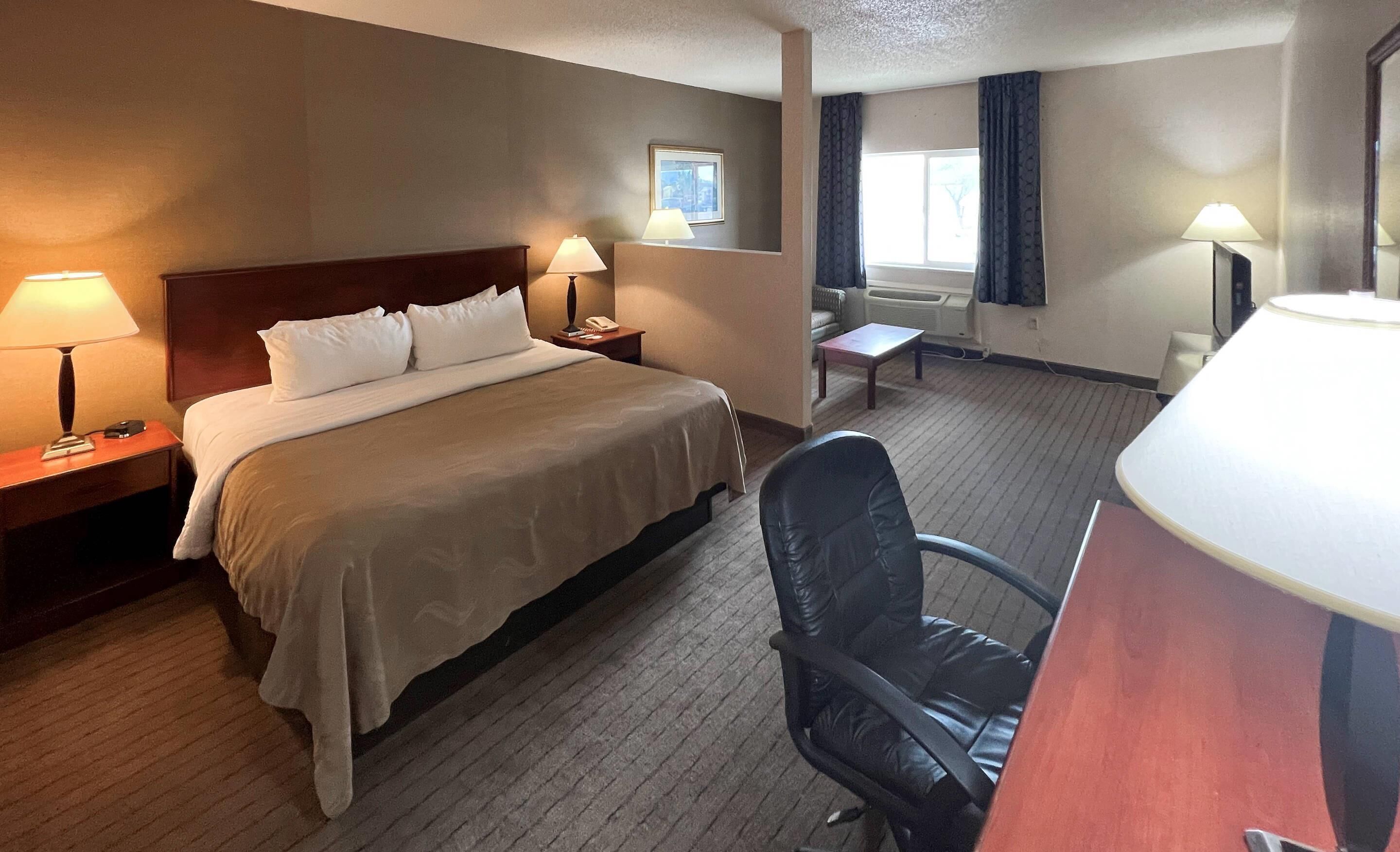 A hotel room with a bed, two bedside tables with lamps, a chair, a small table, and a window with curtains.