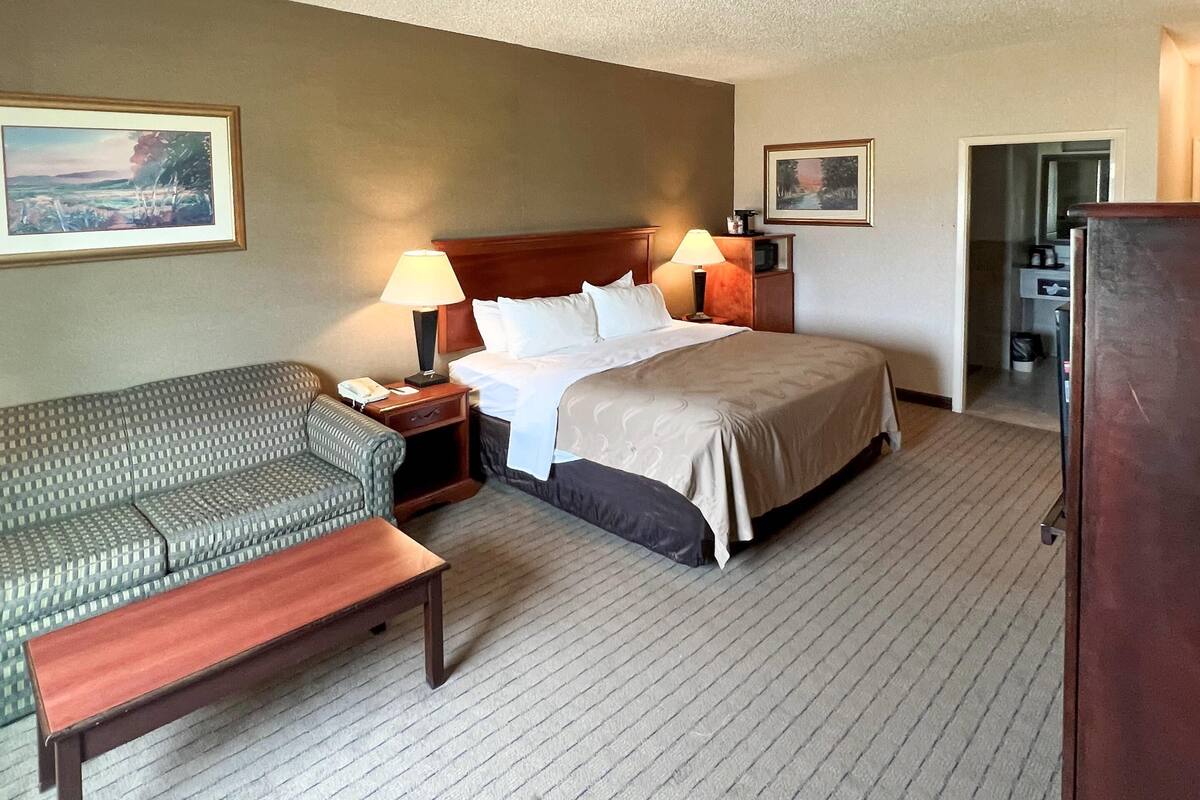 A hotel room with a bed, a sofa, a nightstand, a lamp, and a picture frame on the wall.
