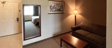 A hotel room with a wooden floor, a sofa, a table, a lamp, a mirror, and a door.