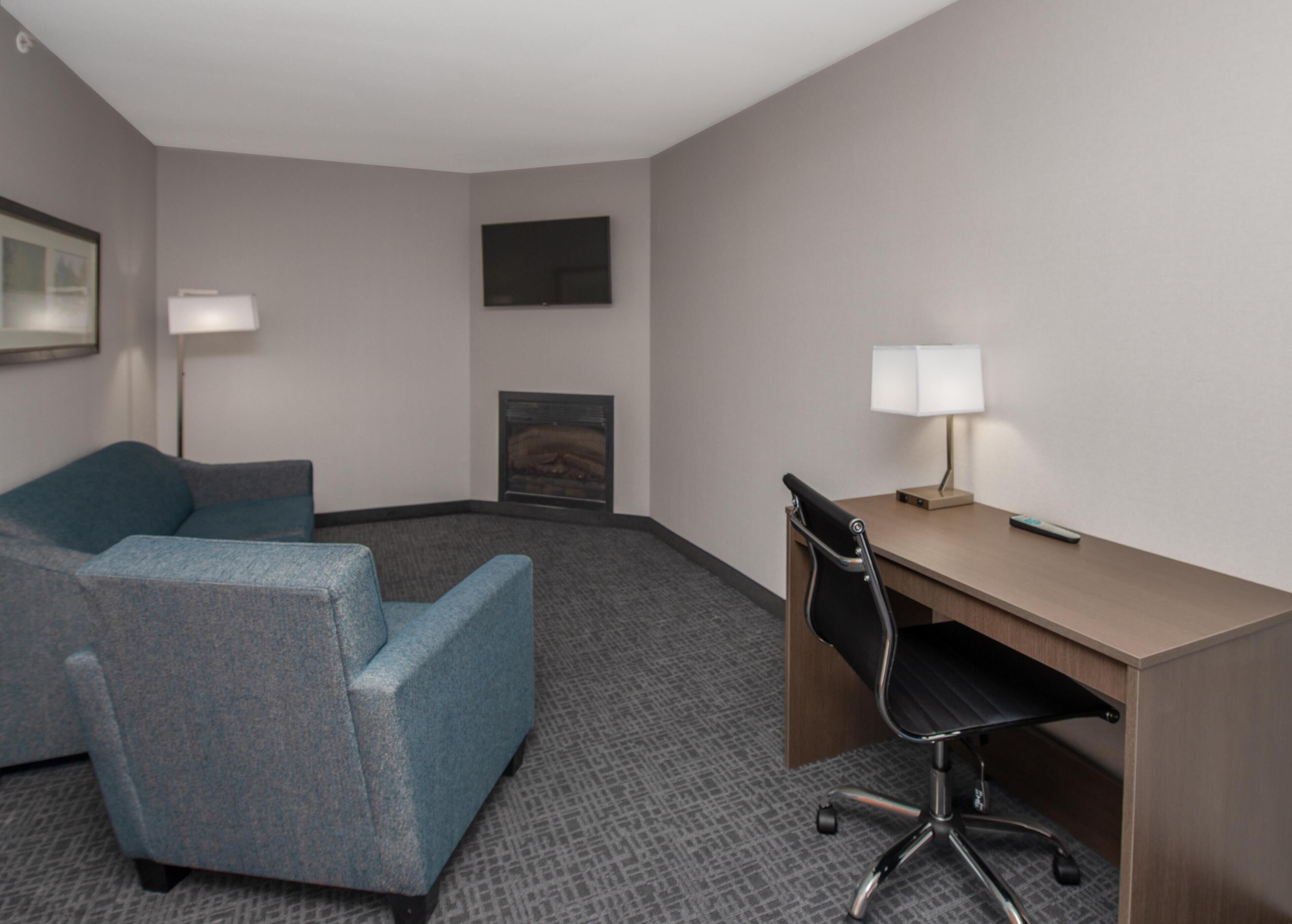 Ein Hotelzimmer mit einem blauen Sofa, einem Holztisch, einem schwarzen Bürostuhl, einem Flachbildfernseher, einem Kamin und einer Lampe.